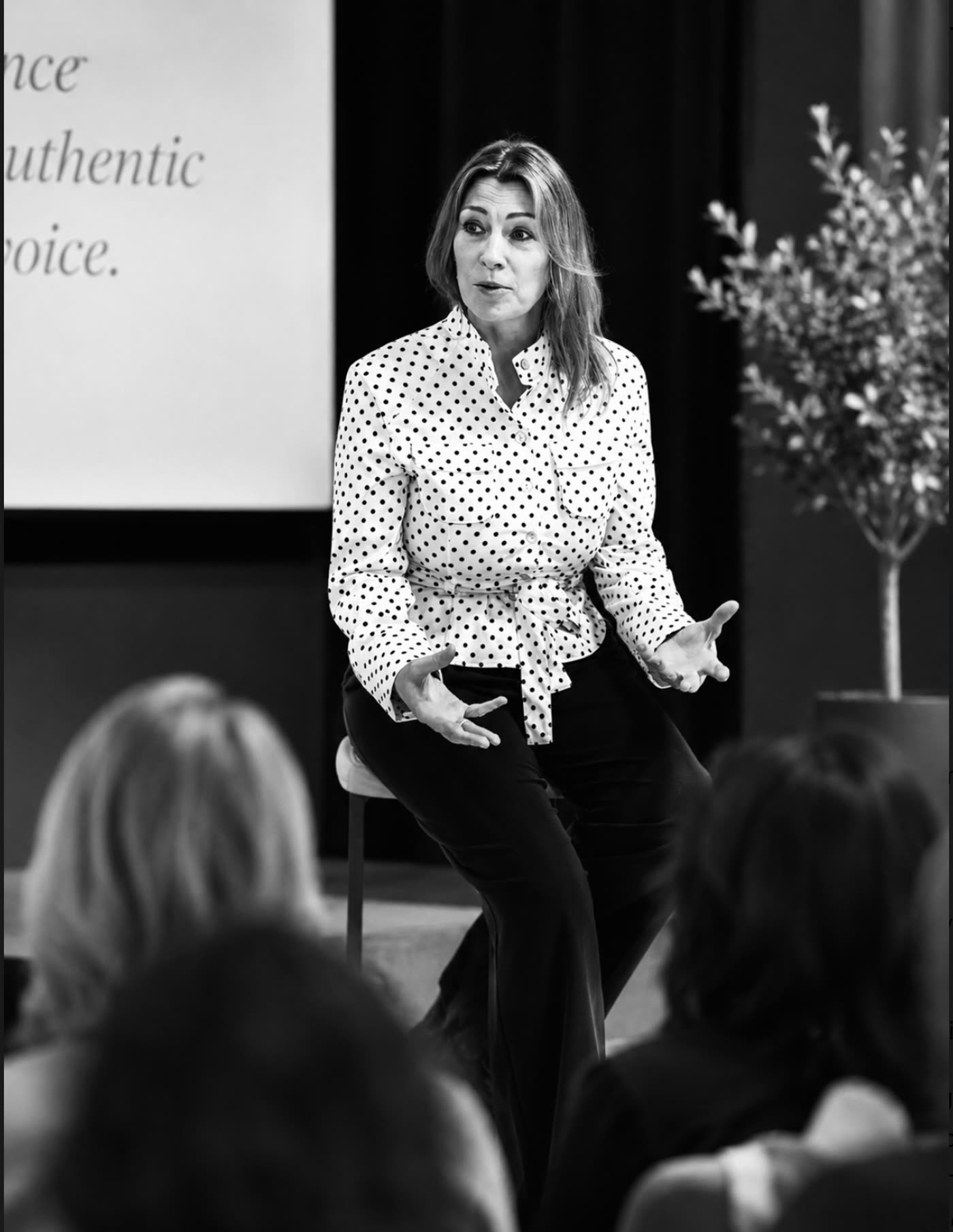 Kate Whelan speaking to an audience, seated on a stool mid-gesture, in black and white.
