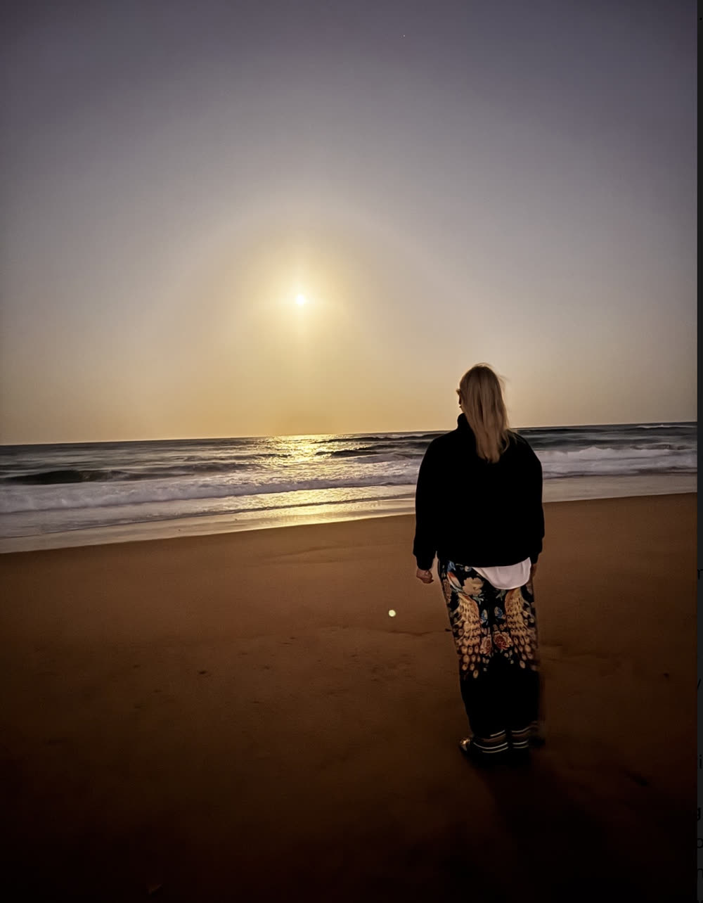 Kate Whelan standing on a beach at dusk, facing the sea and a low sun.